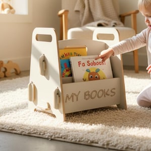 Puede incluir: Un estante para libros de madera de color claro con dos asas superiores y el texto MY BOOKS grabado en el panel frontal. El estante contiene dos libros infantiles, uno con cubierta amarilla y otro con cubierta blanca con una ilustración de dibujos.