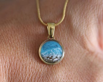 Può includere: Un ciondolo circolare color oro con un inserto in resina in miniatura raffigurante una scena di spiaggia. Il design mostra una riva sabbiosa con ciottoli bianchi che sfuma in un'acqua blu turchese con onde di schiuma bianca. Il ciondolo pende da una catenina.
