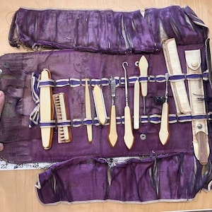 May include: A vintage purple velvet roll-up case with various ivory-colored medical tools. The case is secured with blue ribbon and features a variety of instruments, including a small comb, a knife, and several other tools. The case is open, displaying the tools.