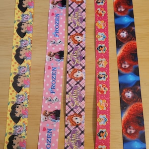 May include: Five different colorful ribbon rolls with cartoon characters printed on them. The ribbons feature characters from Dora the Explorer, Frozen, Sofia the First, Disney Princesses, and Tangled.