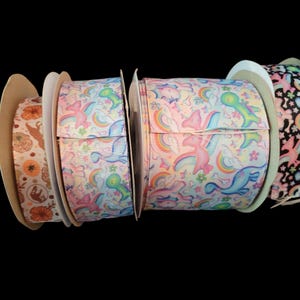 May include: Spools of decorative ribbon in various patterns. One spool features pumpkins and fall leaves. Other spools display unicorns, rainbows, and dinosaurs on a white background. Another has a black background with colorful designs.