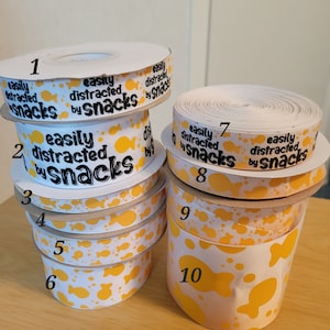 May include: Ten rolls of white ribbon with a yellow and orange print of fish and the text "easily distracted by snacks". The rolls are numbered 1 through 10.