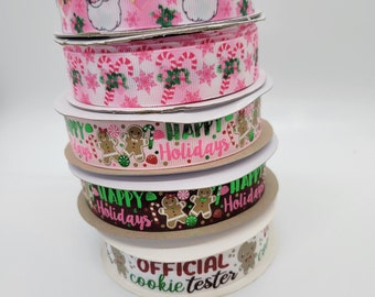 May include: A stack of five rolls of Christmas ribbon. The top ribbon is pink with a white Santa Claus pattern. The second ribbon is pink with a white candy cane and snowflake pattern. The third ribbon is brown with a gingerbread man pattern and the text "Happy Holidays". The fourth ribbon is brown with a gingerbread man pattern and the text "Happy Holidays". The bottom ribbon is white with a gingerbread man pattern and the text "Official Cookie Tester".