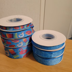 May include: Two spools of blue ribbon with different patterns. One spool has a blue and white pattern with a water ripple effect. The other spool has a blue background with colorful images of flamingos, donuts, and pool floats.