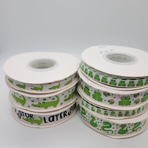 May include: Two spools of white ribbon with green alligator and frog designs. The alligator ribbon has the text "Gator Later" printed on it.