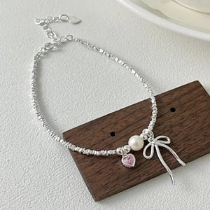 Puede incluir: Una pulsera plateada con una cadena texturizada. La pulsera presenta una perla blanca, un dije de corazón rosa y un dije de lazo. La pulsera se exhibe sobre una superficie de madera.