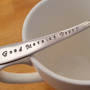 May include: A silver-coloured spoon engraved with "Good Morning Poppy". The spoon is placed over a white mug. The background shows a wooden surface.