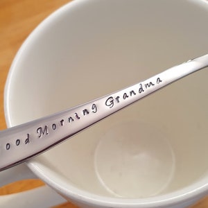 May include: A silver spoon with the words "Good Morning Grandma" engraved on it. The spoon is resting in a white ceramic mug.