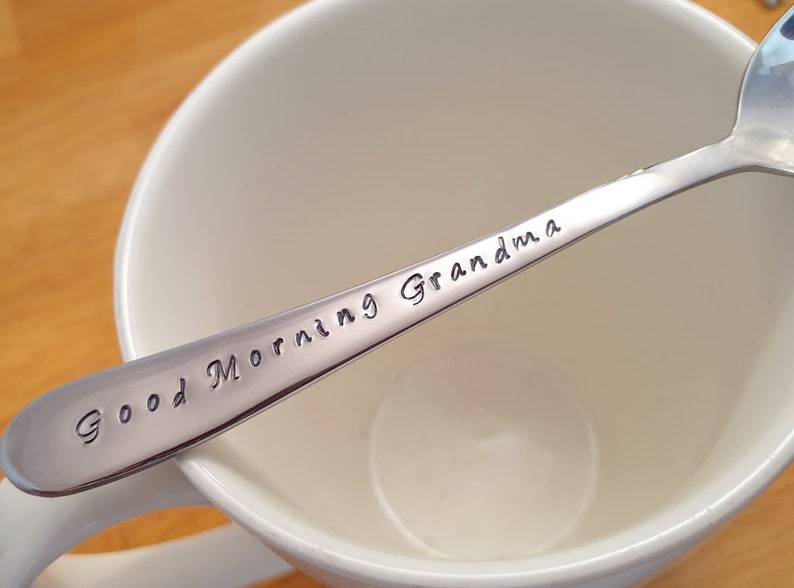 May include: A silver spoon with the words "Good Morning Grandma" engraved on it. The spoon is resting in a white ceramic mug.