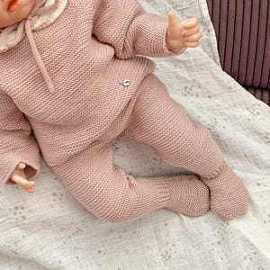 Puede incluir: Una muñeca bebé vestida con un conjunto rosa tejido, que incluye un gorro, un suéter y un pantalón. La muñeca está acostada sobre una almohada y una manta con estampado blanco y rosa. El atuendo tiene un cuello blanco y un pequeño emblema plateado.