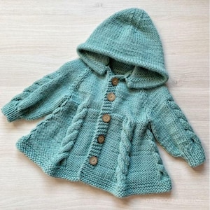 Può includere: Una giacca con cappuccio lavorata a maglia color turchese con bottoni in legno. La giacca presenta dettagli a treccia sul davanti e sulle maniche. Il cappuccio è attaccato al colletto. La giacca è progettata per un bambino.