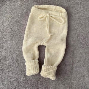 May include: Cream-colored knitted baby pants with a drawstring waist and ribbed cuffs. The soft, textured fabric suggests a cozy and comfortable design. These pants are likely designed for newborns or infants.