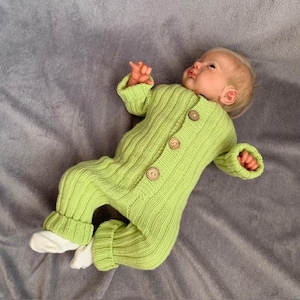 Puede incluir: Un mono de bebé de punto verde claro con botones de madera. El mono tiene mangas y perneras largas con puños acanalados. El bebé lleva calcetines blancos. El conjunto está sobre una superficie gris.