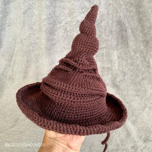 Może przedstawiać: Bordowy kapelusz szydełkowy z szerokim rondem i wysokim, spiralnym czubkiem. Kapelusz jest ręcznie robiony z widocznymi szwami i teksturowaną powierzchnią. Rondo jest okrągłe, a z boku zwisa sznurek. Kapelusz jest w ciemnym odcieniu czerwieni.