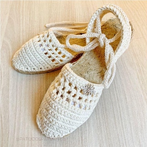 Puede incluir: Un par de sandalias de esparto color crema con un diseño de ganchillo tejido. Las sandalias tienen suela de yute y cintas en el tobillo. Una pequeña flor plateada adorna la parte delantera de una sandalia. Las sandalias se exhiben sobre una superficie de madera clara.