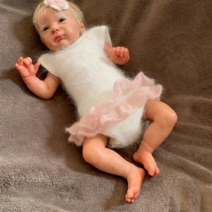Puede incluir: Una muñeca bebé realista que lleva un body blanco de punto con una falda rosa con volantes. La muñeca tiene el pelo rubio, una diadema rosa y está tumbada sobre una superficie marrón. La piel de la muñeca tiene un tono realista.