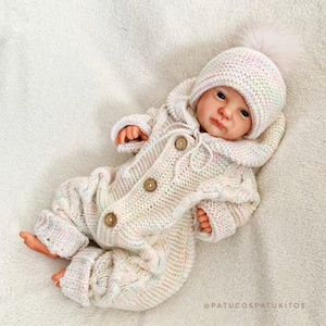May include: A baby doll wearing a cream-colored knitted outfit with a matching hat. The outfit has a hooded jacket with wooden buttons and a cable-knit design. The hat is topped with a fluffy pom-pom.