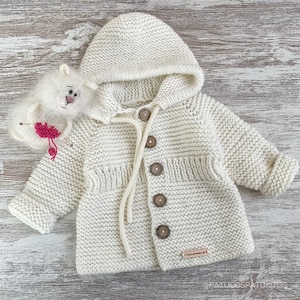 Puede incluir: Chaqueta de bebé de punto color crema con capucha y botones de madera. La chaqueta tiene un patrón de punto texturizado y una pequeña etiqueta. Un peluche blanco con detalles rosas está al lado de la chaqueta.