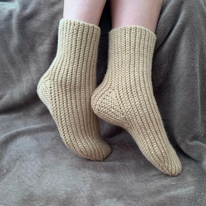 Puede incluir: Un par de calcetines beige tejidos. Los calcetines son acanalados y parecen hechos a mano. Los calcetines están sobre una superficie gris.