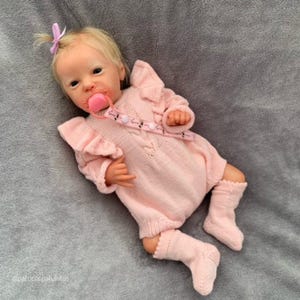 May include: A lifelike baby doll with blonde hair and a pink bow. The doll wears a pink knitted romper with ruffled sleeves and matching socks. A pink pacifier is in its mouth, and a pacifier clip is attached.