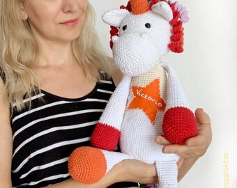 May include: A handmade white crochet unicorn plush toy with a bright orange horn, red and orange accents, and a star with the name "Victoria". The unicorn has a colorful mane and is being held.