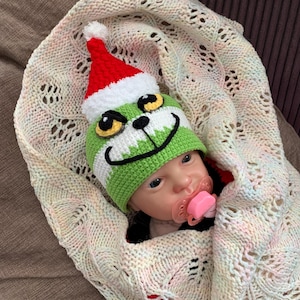 May include: A baby wearing a green and white crocheted hat with a Grinch face and a red Santa hat. The baby is wrapped in a cream-colored knitted blanket with a pink pacifier in its mouth.