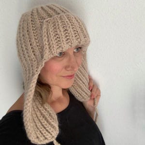 May include: A beige knitted hat with ear flaps. The hat is made of a thick, ribbed knit material. The ear flaps extend down and are tied with a string. The hat covers the head and ears, providing warmth and protection from the cold.