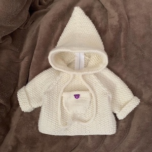 Op de afbeelding: Een crèmekleurig, gebreid babyjasje met een capuchon. De jas heeft een voorzak met een klein paars berenontwerp. De mouwen zijn omgeslagen en de capuchon is puntig. De jas is gemaakt van een getextureerde, dikke brei.