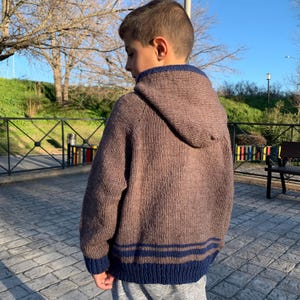May include: A brown hooded sweater with navy blue trim and stripes. The sweater is knitted and has a full zipper. The hood is lined with navy blue fabric. The sweater is worn by a person.