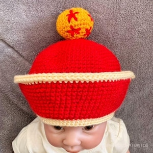 Puede incluir: Un gorro de bebé tejido a ganchillo rojo y amarillo. El gorro es principalmente rojo con un borde amarillo y una bola amarilla en la parte superior con detalles en forma de estrella roja. El gorro está diseñado para la cabeza de un bebé.