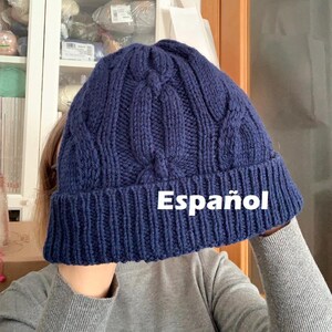 May include: A navy blue knitted beanie hat with a cable knit design. The hat is made of a thick, textured yarn and has a folded brim. The word "Español" is printed on the hat.