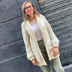 May include: Cream-colored, open-front cardigan sweater. The sweater has a shawl collar and long sleeves. The person is wearing a white top and olive green pants. The background is a gray brick wall and a gray metal door.