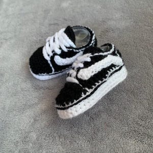 May include: A pair of handmade baby trainers in black and white crochet. The miniature shoes feature a classic trainer design with white laces, a white sole, and black accents. The shoes are displayed on a grey surface.