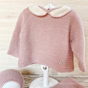 Peut inclure: Un pull rose clair en tricot avec un col Claudine crème. Le pull a des manches longues et un petit emblème argenté près de l'ourlet. Un bonnet et une couverture assortis sont également visibles.
