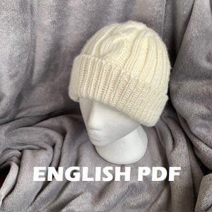 Puede incluir: Gorro de punto color crema con borde acanalado y diseño de punto trenzado texturizado. El gorro se muestra sobre una cabeza de maniquí blanca sobre un fondo gris texturizado. El texto "ENGLISH PDF" es visible.