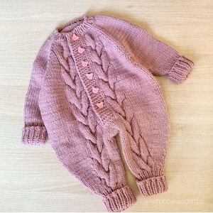 May include: A knitted, mauve-colored baby romper with a cable knit design. The romper features long sleeves, ribbed cuffs, and a button-up front with pink bear-shaped buttons. The garment is designed for infants.