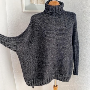 Puede incluir: Jersey de cuello alto gris oscuro de gran tamaño con cuello, puños y dobladillo de canalé. El jersey tiene un ajuste holgado y cómodo con mangas largas. La prenda de punto se exhibe sobre una forma de madera sobre un fondo neutro.