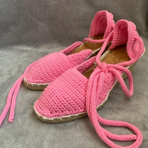 May include: A pair of pink crochet espadrille sandals. The sandals have a woven jute sole, a closed toe, and ankle ties. The crochet work is a solid pink color, and the soles are trimmed with white stitching.