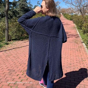 Könnte beinhalten: Ein marineblauer Strickcardigan mit langem, offenem Design. Der Cardigan hat ein strukturiertes Stichmuster und einen geteilten Rücken. Die Ärmel sind lang und der Stil ist entspannt. Der Cardigan wird im Freien auf einem Ziegelweg getragen.