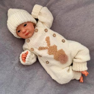 May include: A cream-colored knitted baby outfit with a matching hat. The outfit features a brown bunny design on the front and wooden buttons. A small, pink pom-pom serves as the bunny's tail. The baby doll is lying on a gray surface.