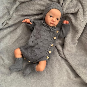 Puede incluir: Una muñeca bebé realista vestida con un conjunto de punto gris, que incluye un gorro, un cárdigan con botones y calcetines. El conjunto tiene un patrón texturizado y botones de madera. La muñeca está sobre una manta gris suave.