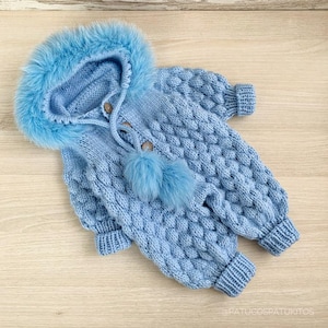 Puede incluir: Traje de nieve de bebé de punto azul claro con capucha ribeteada de piel sintética. El traje de nieve presenta un patrón de punto de cable, botones de madera y dos pompones esponjosos. Los puños y los tobillos son acanalados.