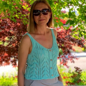 May include: A light turquoise knitted tank top with a button-up front. The top has a scoop neck and a delicate openwork pattern. The buttons are a light brown color. The background features green and red foliage.