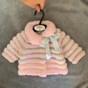 Puede incluir: Una chaqueta de bebé rosa y a rayas con cuello esponjoso y lazo azul claro. La chaqueta tiene rayas horizontales rosas, verdes y amarillas. La percha tiene una etiqueta que dice "IRINA Emelina".