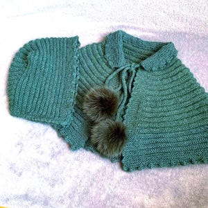 Puede incluir: Un conjunto de capa y gorro de punto color verde azulado. La capa presenta un diseño acanalado, un cuello y un cierre de lazo. Dos pompones esponjosos de color marrón oscuro están unidos en la parte delantera. El gorro tiene un patrón acanalado similar.