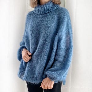 Puede incluir: Un jersey de cuello alto azul claro de gran tamaño. El jersey tiene un ajuste holgado y cómodo con mangas largas. La prenda de punto parece suave y esponjosa, perfecta para climas más fríos. El jersey es de color sólido, con un cuello alto y acanalado.