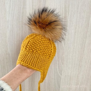 Puede incluir: Gorro de punto amarillo mostaza con orejeras y un gran pompón marrón y negro esponjoso en la parte superior. El gorro tiene un patrón de punto texturizado y lazos largos. Fondo de madera clara.