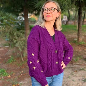May include: A deep purple cardigan with a V-neck and cable knit design. The sleeves are adorned with star-shaped embroidery, and the cardigan fastens with two brown buttons. The garment is worn with blue jeans.