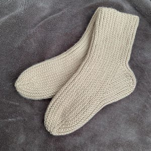 Puede incluir: Un par de calcetines beige tejidos, colocados sobre una superficie gris texturizada. Los calcetines presentan un patrón de punto acanalado, lo que indica una fabricación artesanal. El color neutro y el diseño sencillo sugieren comodidad y versatilidad.