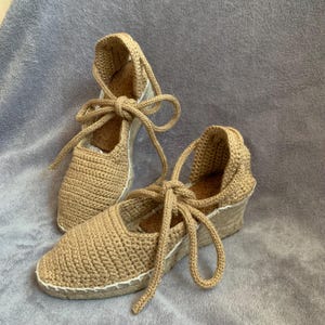 Op de afbeelding: Een paar bruin gehaakte espadrille sandalen met sleehakken. De sandalen hebben een dichte neus, enkelbanden en een wit gestikte zool. Het haakmateriaal geeft een textuur.